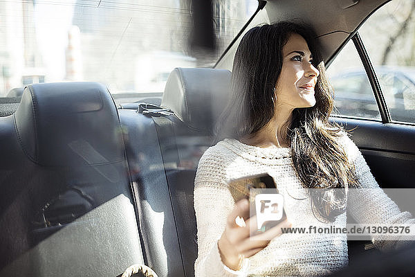 Nachdenkliche Frau hält Smartphone in der Hand  während sie durch ein Taxifenster schaut