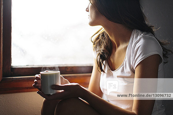 Midsection of Woman with cup sitting by window at home