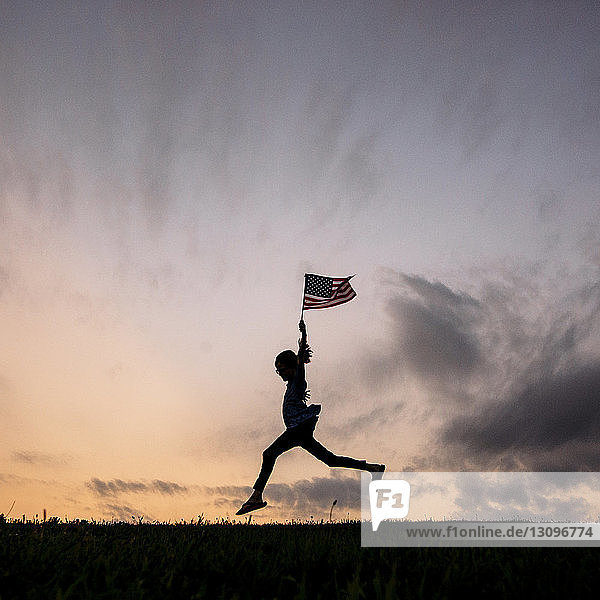 Silhouettenmädchen mit amerikanischer Flagge läuft auf Feld gegen Himmel