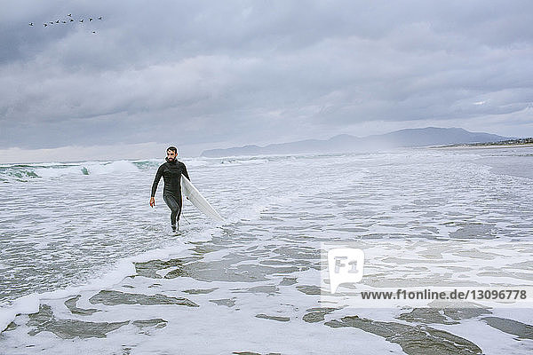 Mann mit Surfbrett schaut weg  während er im Meer gegen bewölkten Himmel läuft