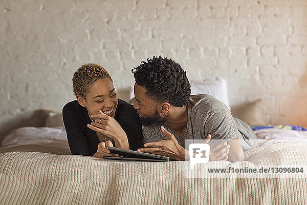 Glückliches Paar unterhält sich  während es zu Hause am Bett einen Tablet-Computer benutzt