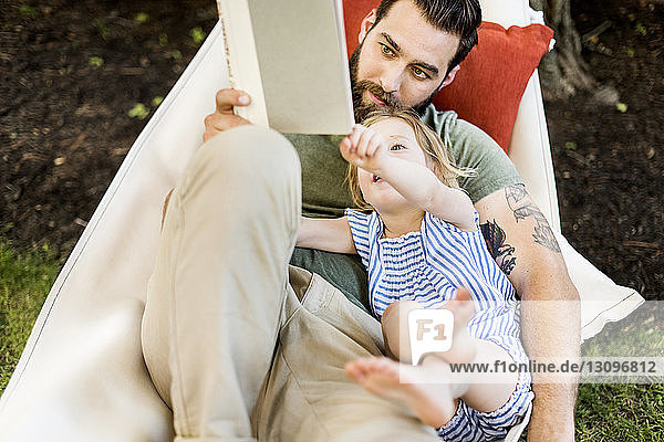Hochwinkelansicht einer Tochter mit einem Vater  der ein Buch liest  während sie auf einer Hängematte im Hof liegt