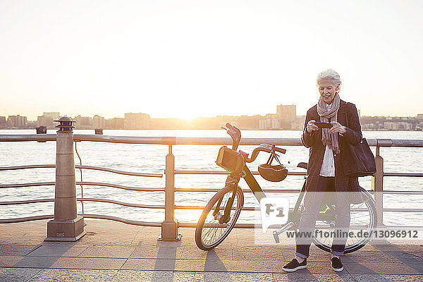 Ältere Frau benutzt Smartphone mit dem Fahrrad auf Promenade gegen klaren Himmel