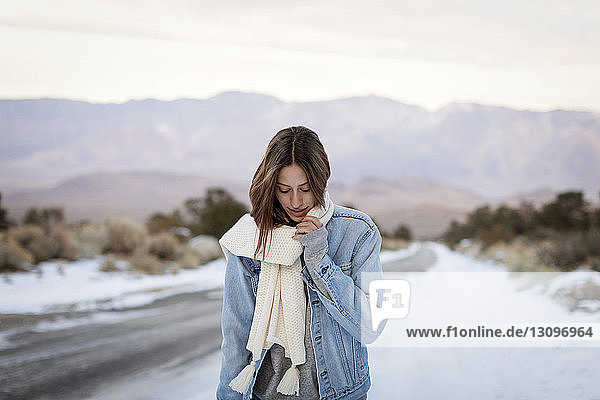 Young woman holding muffler while standing on snow covered road against mountains