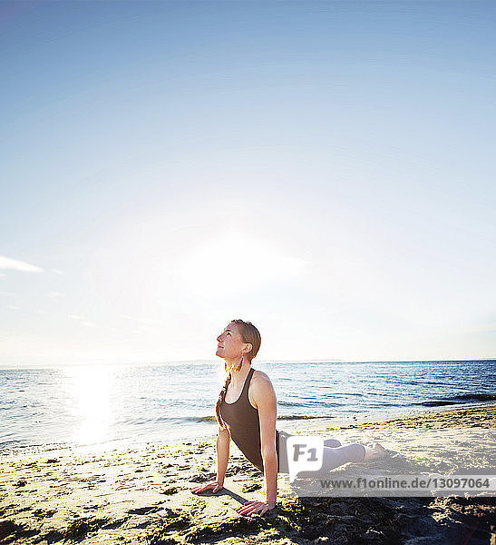 Woman practicing cobra pose at beach against sky during sunny day