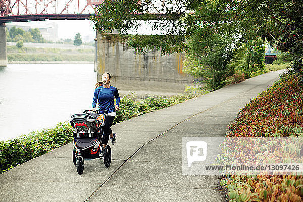 Frau joggt mit Kinderwagen auf einem Fussweg am Fluss