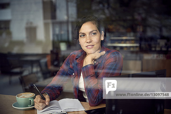Nachdenkliche Frau mit Buch und Cappuccino auf dem Tisch sitzend im Cafe durchs Fenster gesehen