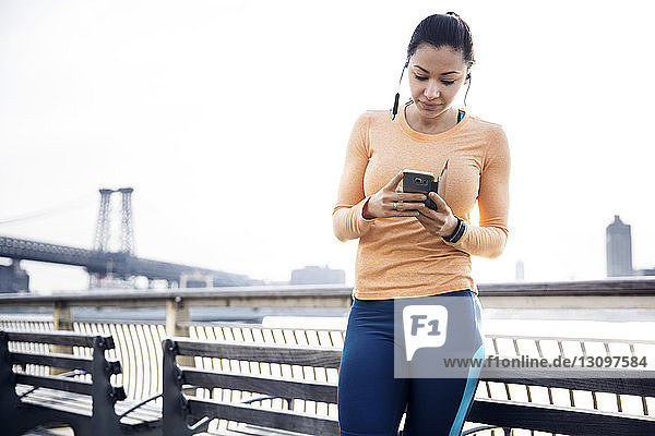 Sportliche Frau hört Musik über Smartphone mit Williamsburg Bridge im Hintergrund