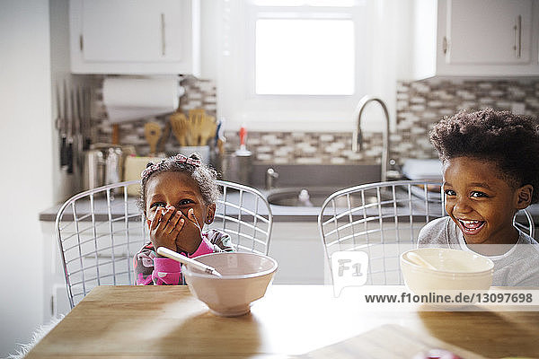 Cheerful brother and sister looking away while having breakfast in kitchen
