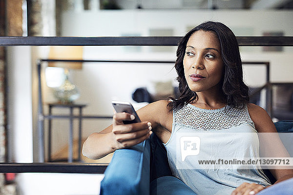 Nachdenkliche Geschäftsfrau mit Smartphone schaut weg  während sie im Kreativbüro auf dem Sofa sitzt