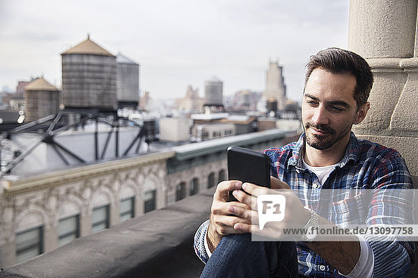 Lächelnder Mann benutzt Smartphone  während er auf dem Balkon gegen den Himmel sitzt