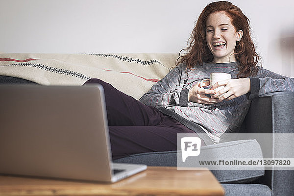 Cheerful woman with cup looking at laptop computer while sitting on sofa at home
