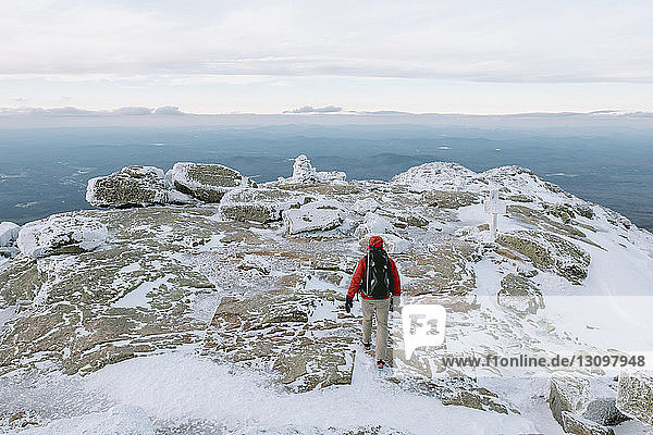 Rückansicht eines Rucksacktouristen mit Kamera beim Gehen auf schneebedeckten Felsen