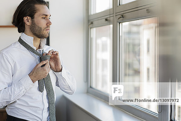 Thoughtful man looking through window tying necktie at home