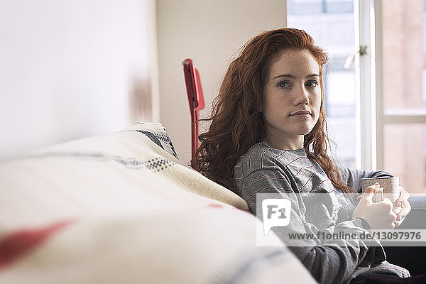 Nachdenkliche Frau hält Kaffeetasse  während sie sich zu Hause auf dem Sofa entspannt