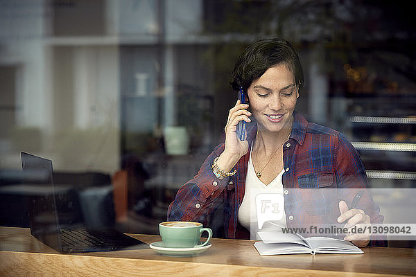 Frau spricht am Smartphone  während sie im Café sitzt und durch das Fenster gesehen wird