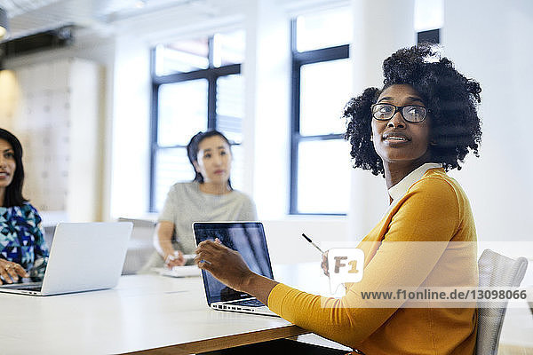 Geschäftsfrau schaut über die Schulter  während sie am Laptop-Computer mit Kolleginnen im Hintergrund arbeitet