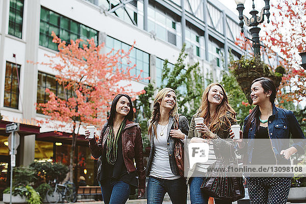 Glückliche Freundinnen halten Kaffeetassen in der Hand und unterhalten sich beim Spaziergang auf der Straße