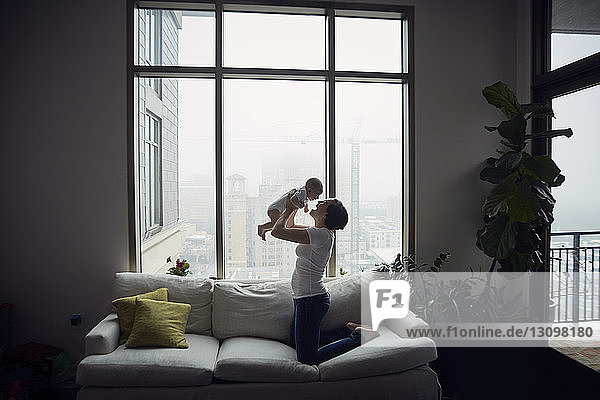 Seitenansicht einer glücklichen Mutter  die mit ihrem Sohn spielt  während sie zu Hause auf dem Sofa am Fenster kniet