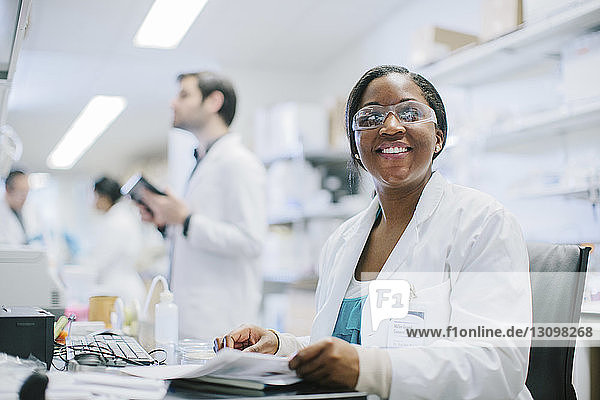 Porträt einer lächelnden Ärztin  die am Schreibtisch sitzt  während ihre Kollegen im Behandlungszimmer arbeiten