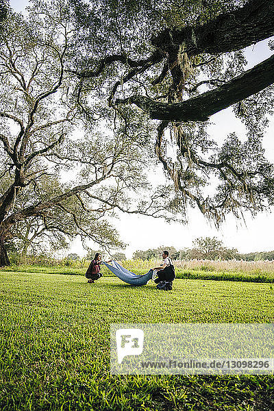 Side view of couple placing blanket on grassy field against sky at park