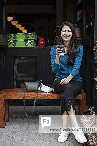 Porträt einer glücklichen Frau  die ein Einwegglas hält  während sie auf einer Bank vor einem Café sitzt