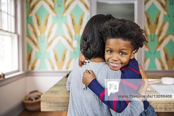 Porträt eines glücklichen Jungen  der seine Mutter umarmt  während er zu Hause Buntstifte hält
