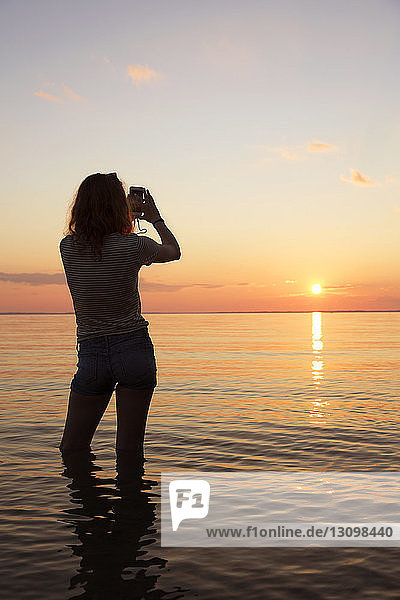 Rückansicht einer Teenagerin  die den Sonnenuntergang fotografiert  während sie im Meer steht