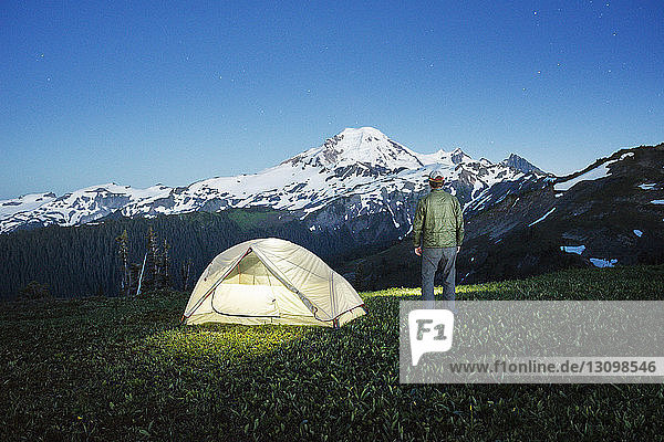 Rear view of man standing by illuminated tent on grassy field against mountain at dusk
