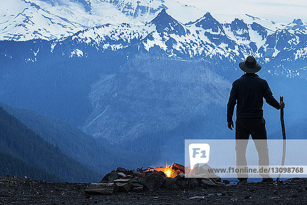 Mann hält Stock am Lagerfeuer gegen schneebedeckte Berge