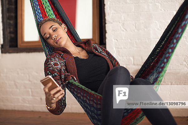 Junge Frau benutzt Smartphone  während sie sich zu Hause in der Hängematte entspannt