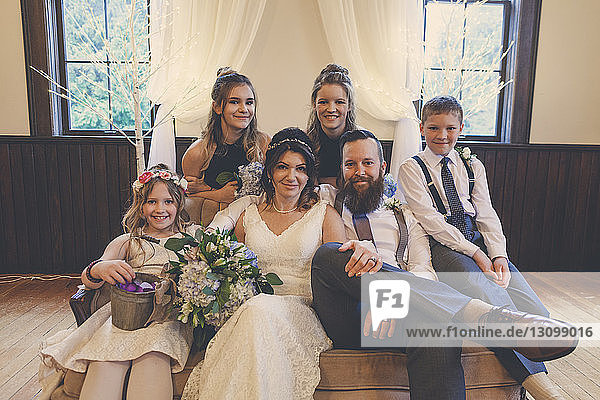 Portrait of newlywed couple sitting by pageboy and flower girl with bridesmaids on sofa during wedding ceremony