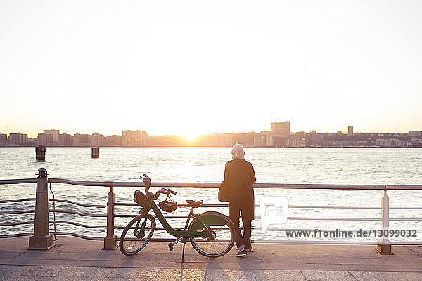 Rückansicht einer älteren Frau  die bei Sonnenuntergang am Fahrrad gegen das Meer steht