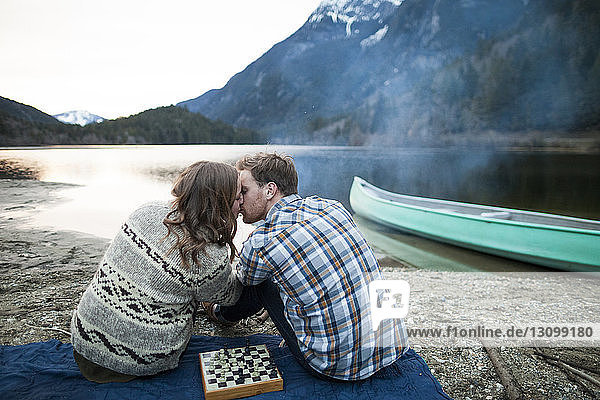 Junges Paar küsst sich beim Sitzen auf einer Picknickdecke am Seeufer