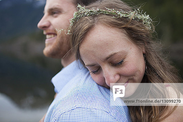 Freundin mit geschlossenen Augen trägt einen Kranz  während sie ihren Freund am Seeufer umarmt
