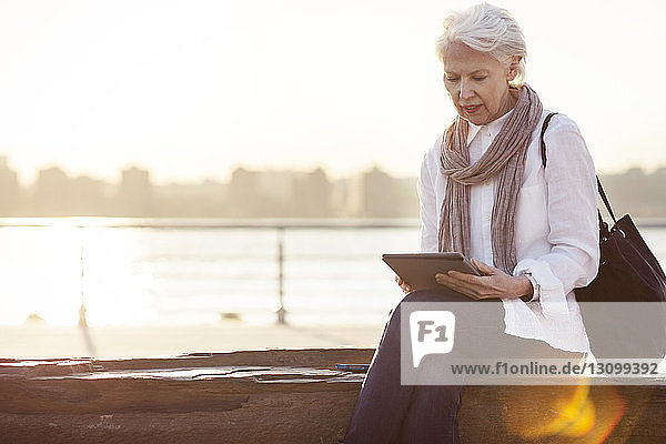 Ältere Frau mit Tablet-Computer auf Stützmauer am Fluss sitzend