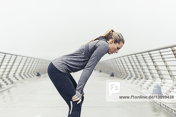 Seitenansicht einer müden Sportlerin  die mit den Händen auf den Knien auf einer Brücke vor klarem Himmel steht