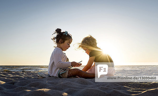 Schwestern sitzen bei Sonnenuntergang auf Sand am Strand vor klarem Himmel