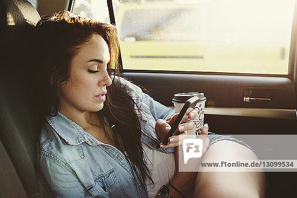 Frau mit Einwegbecher  die während der Fahrt im Auto ein Mobiltelefon benutzt