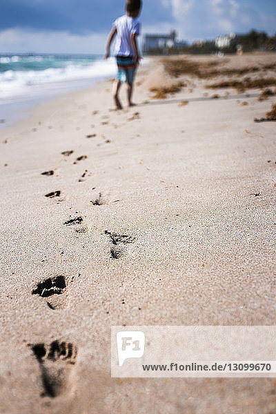 Rückansicht eines Jungen  der beim Laufen auf Sand am Strand Fußspuren hinterlässt
