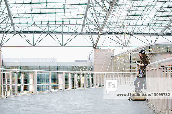 Seitenansicht einer Frau mit Gepäck bei der Benutzung eines Mobiltelefons  während sie sich am Flughafen an die Reling lehnt