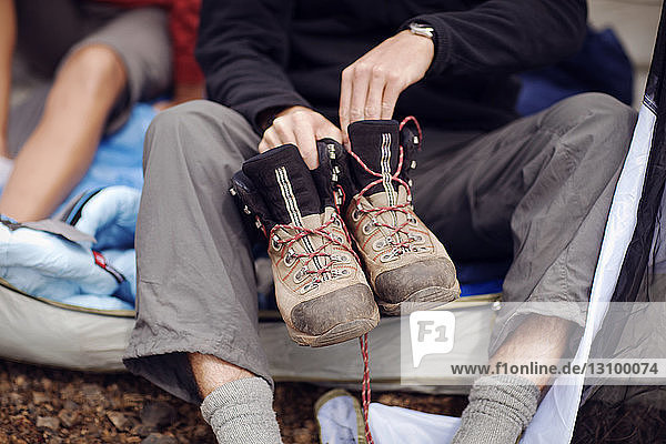Midsection of male hiker holding shoes while sitting in tent at campsite