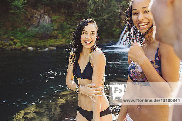 Happy female friends wearing bikini standing at riverbank in forest