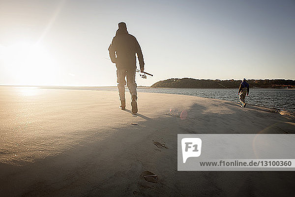 Rückansicht von Männern  die bei Sonnenuntergang mit der Angelrute im Sand gehen