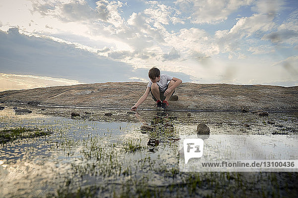 Boy crouching by stream on Arabia Mountain against cloudy sky during sunset
