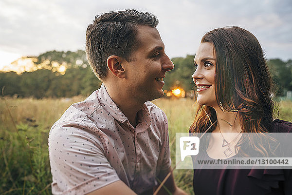 Couple looking at each other while sitting on grassy field against sky