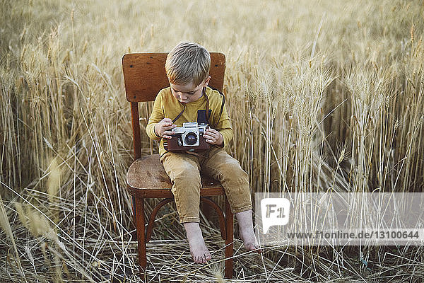 Full length of boy with vintage camera sitting on chair amidst wheat field