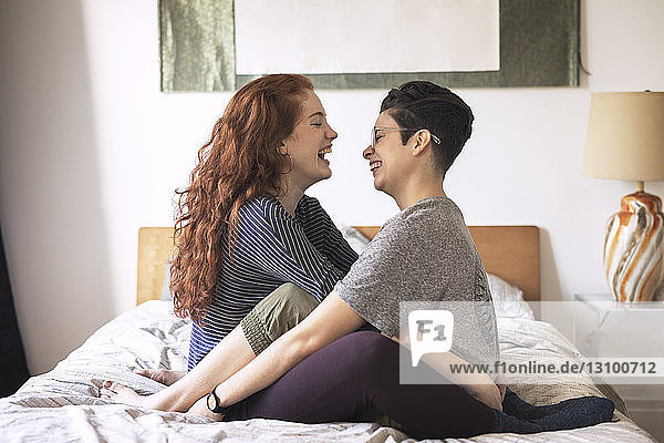 Side view of cheerful lesbians looking at each other while sitting on bed