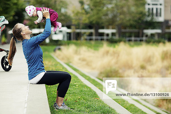 Seitenansicht einer Frau  die ein Baby anhebt  während sie auf einer Stützmauer im Park sitzt
