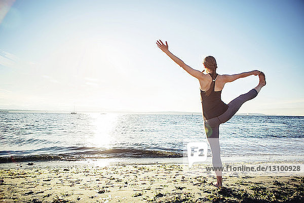 Rückansicht einer Frau  die an einem sonnigen Tag am Strand eine Yogastellung von der Hand bis zum großen Zeh praktiziert